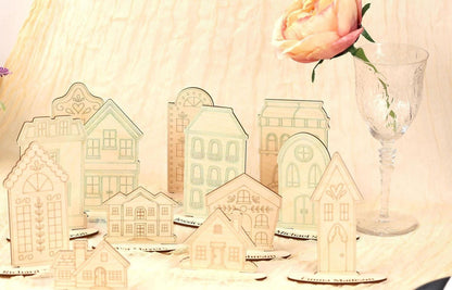 a collection of wooden cutouts of houses, each with unique architectural details, arranged on a table alongside a glass vase with a single pink rose.