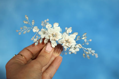 A hand holding a decorative hair accessory with white flowers against a blue background.