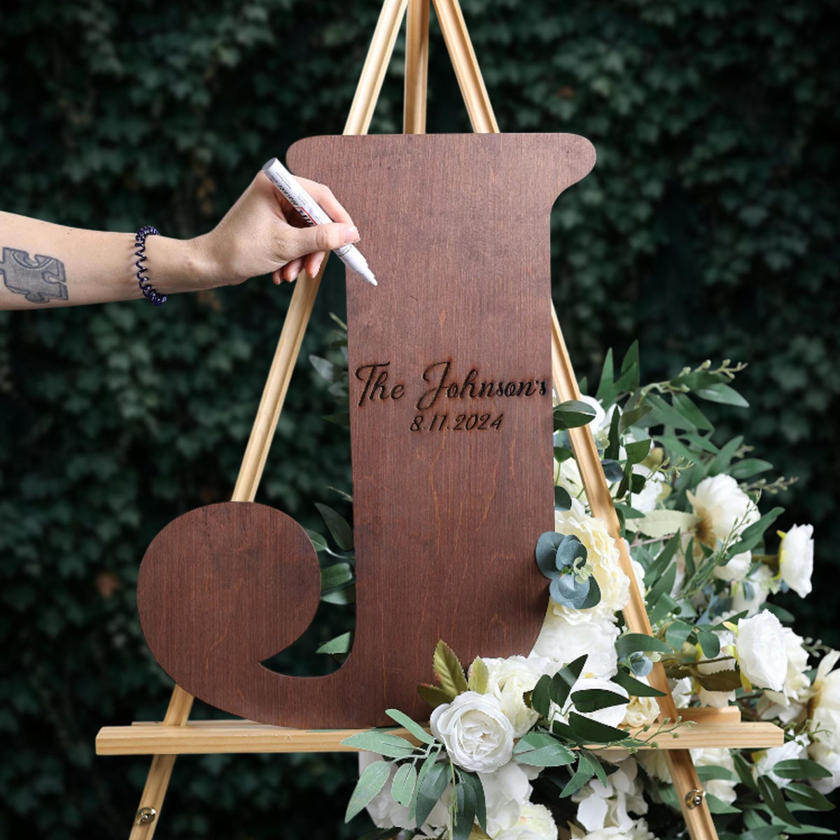 A person is writing on a wooden sign that says "The Johnsons" and "11/11/2024" using a marker.