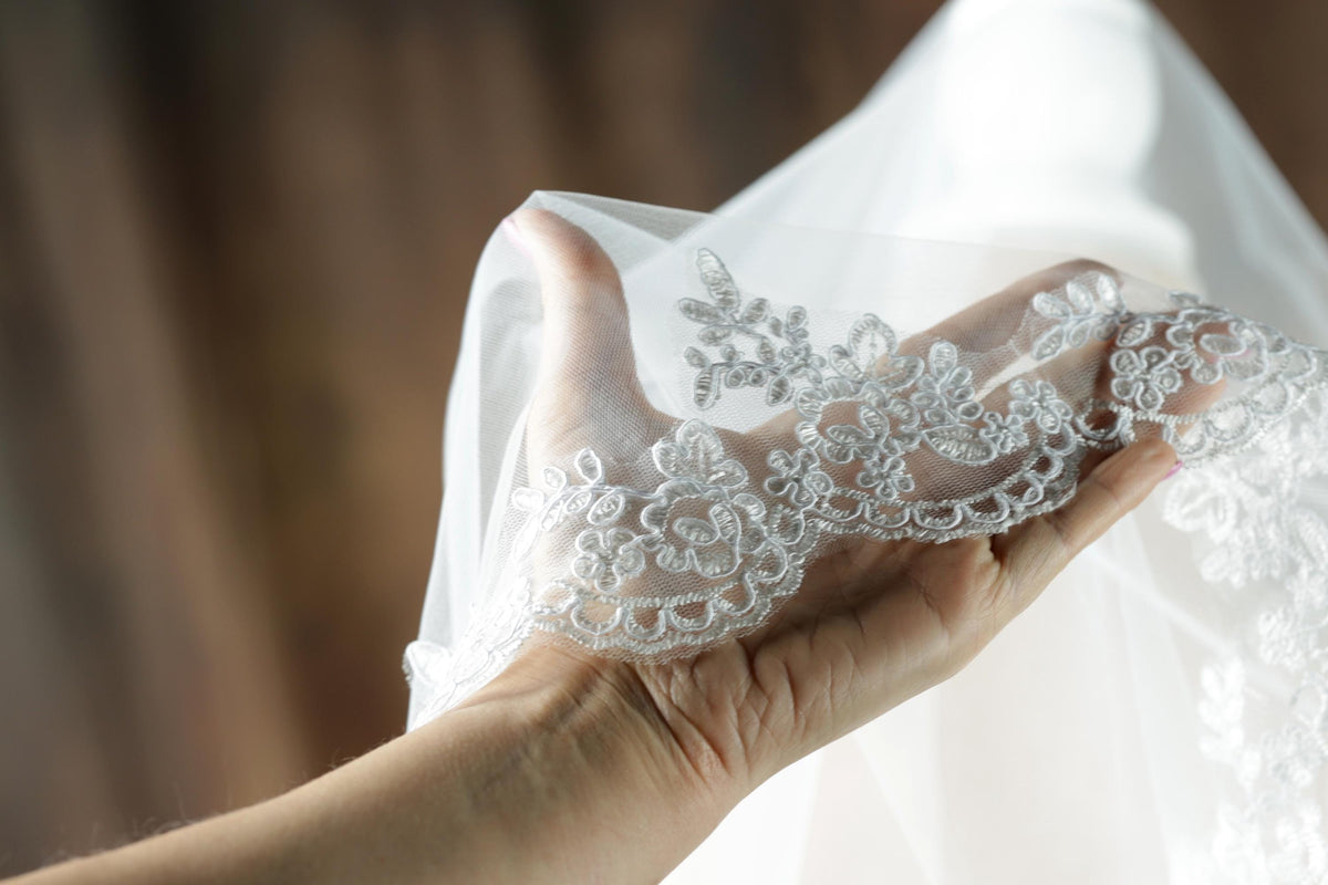 A hand holding a delicate white lace veil with intricate floral patterns.