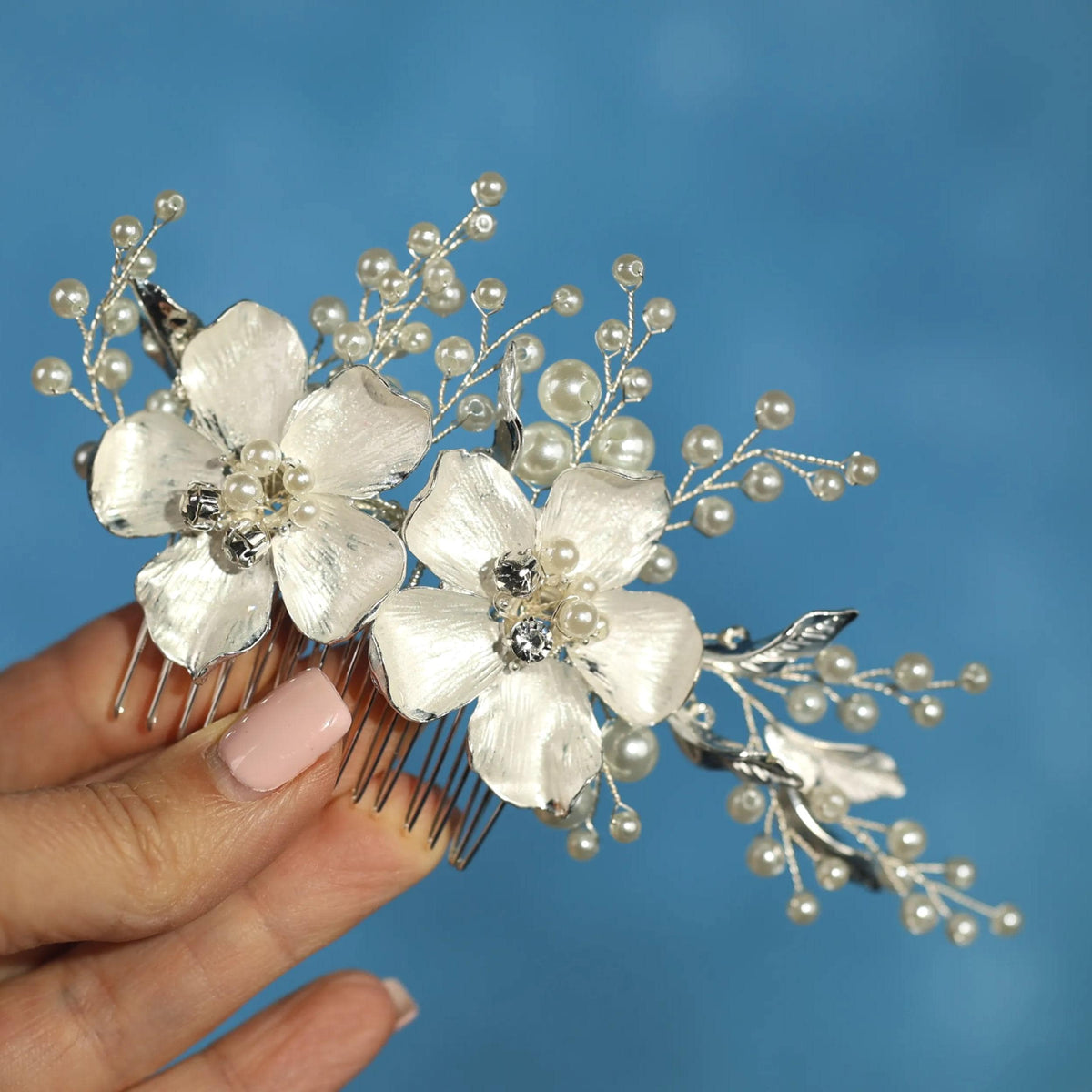A hand holding a decorative hair accessory with white flowers and pearls against a blue background.