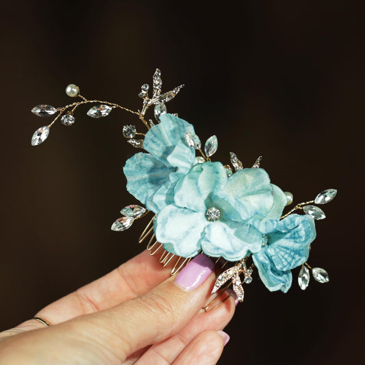 A hand holding a decorative hair accessory with blue and white flowers and sparkling rhinestones.