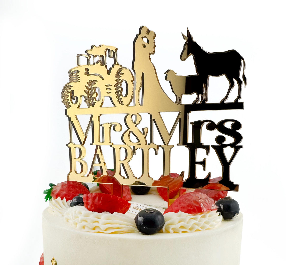 A white cake with a gold and black wedding cake topper featuring a bride and groom silhouette, a tractor, and a donkey.
