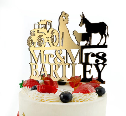 A white cake with a gold and black wedding cake topper featuring a bride and groom silhouette, a tractor, and a donkey.