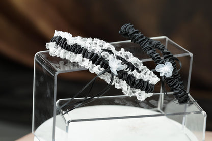 A clear glass display case containing two black and white lace wedding garter belts with white flower accents.