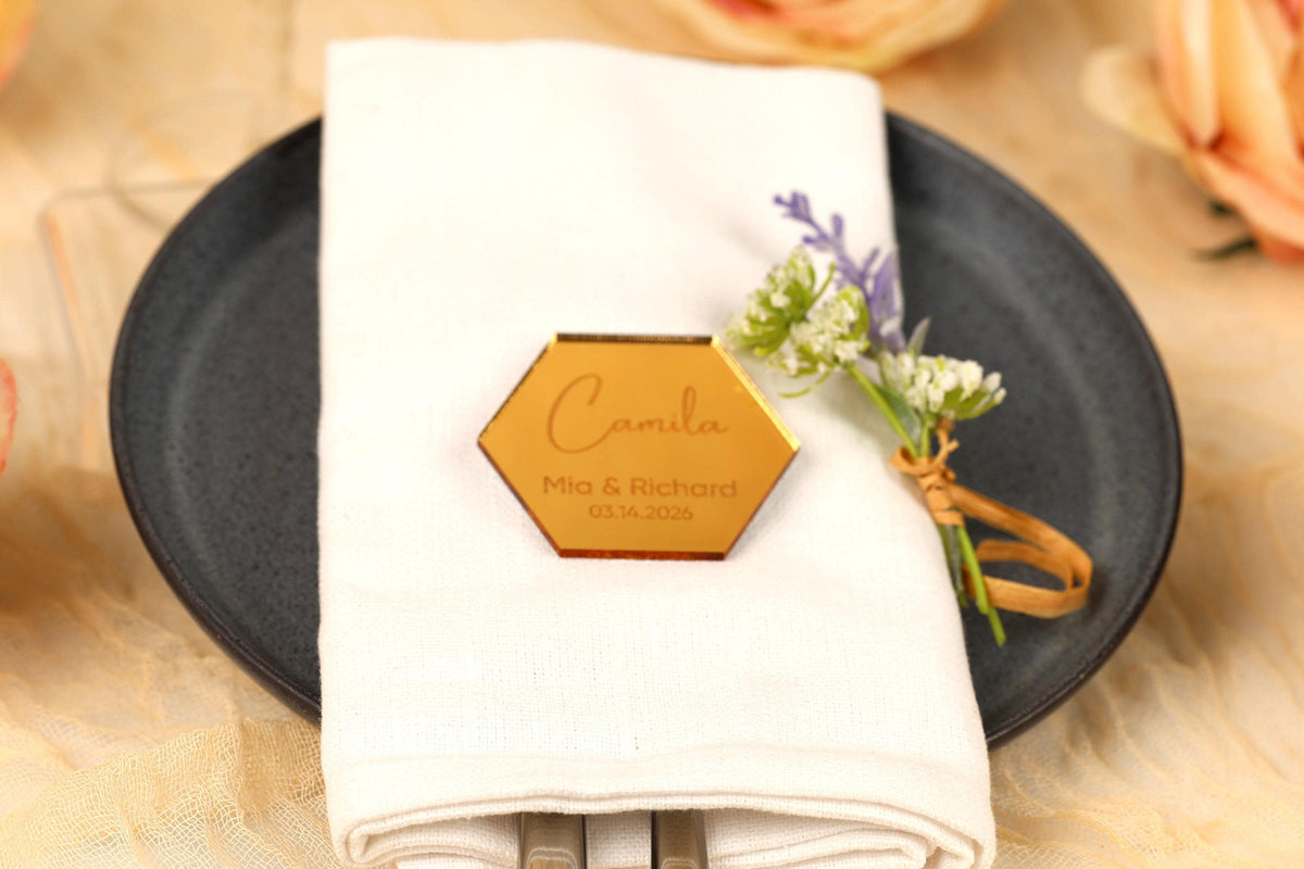 A place setting with a white napkin, a black plate, and a small floral arrangement. There is a hexagonal name tag on the napkin with the names 'Mia & Richard' and the date '10.10.20'.