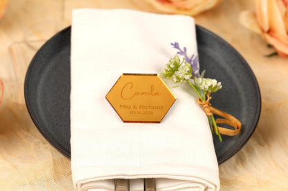 A place setting with a white napkin, a black plate, and a small floral arrangement. There is a hexagonal name tag on the napkin with the names 'Mia & Richard' and the date '10.10.20'.