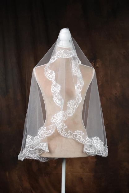 A white lace veil with intricate floral patterns is displayed on a mannequin against a brown background.