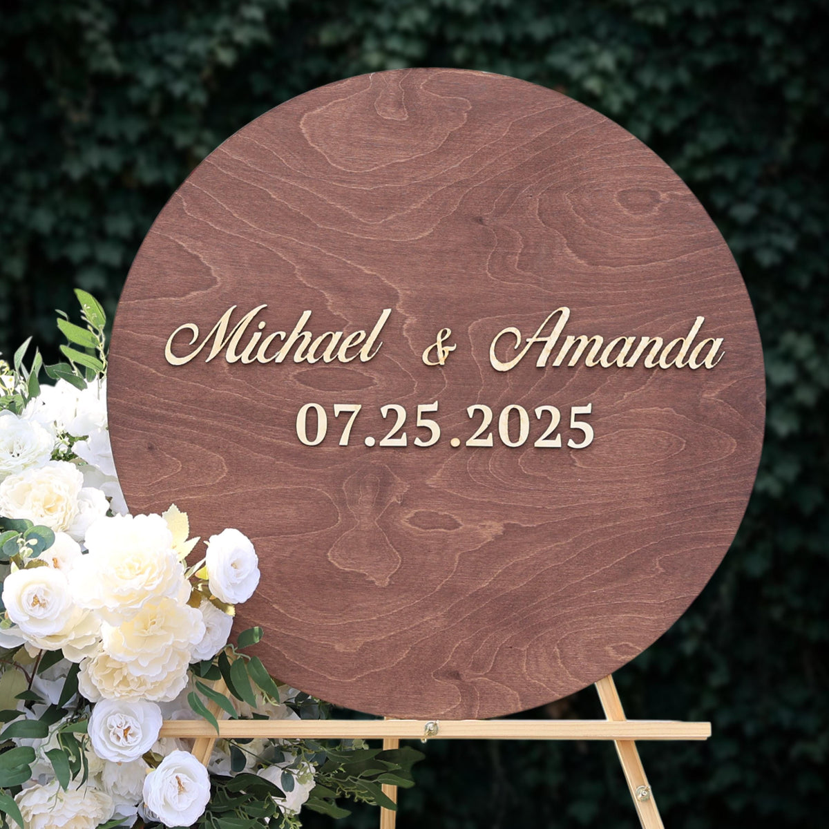 A wooden sign displaying the names "Michael & Amanda" and the date "07.25.2025" is surrounded by a bouquet of white flowers.