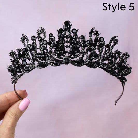 A hand holding a black tiara adorned with numerous black gemstones against a pink background.