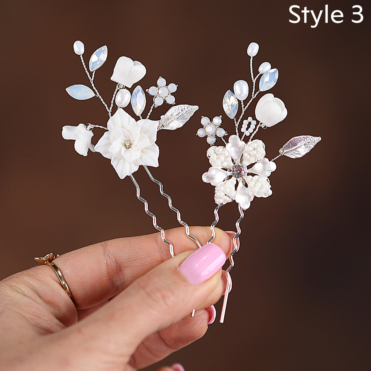 A hand holding two silver hair clips adorned with white flowers and leaves.