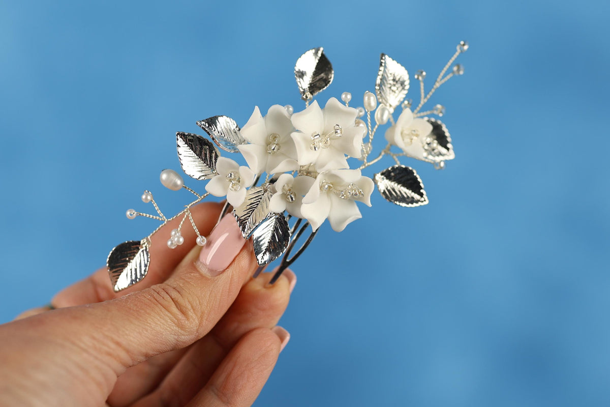 A hand holding a decorative hair clip with white flowers and silver leaves against a blue background.