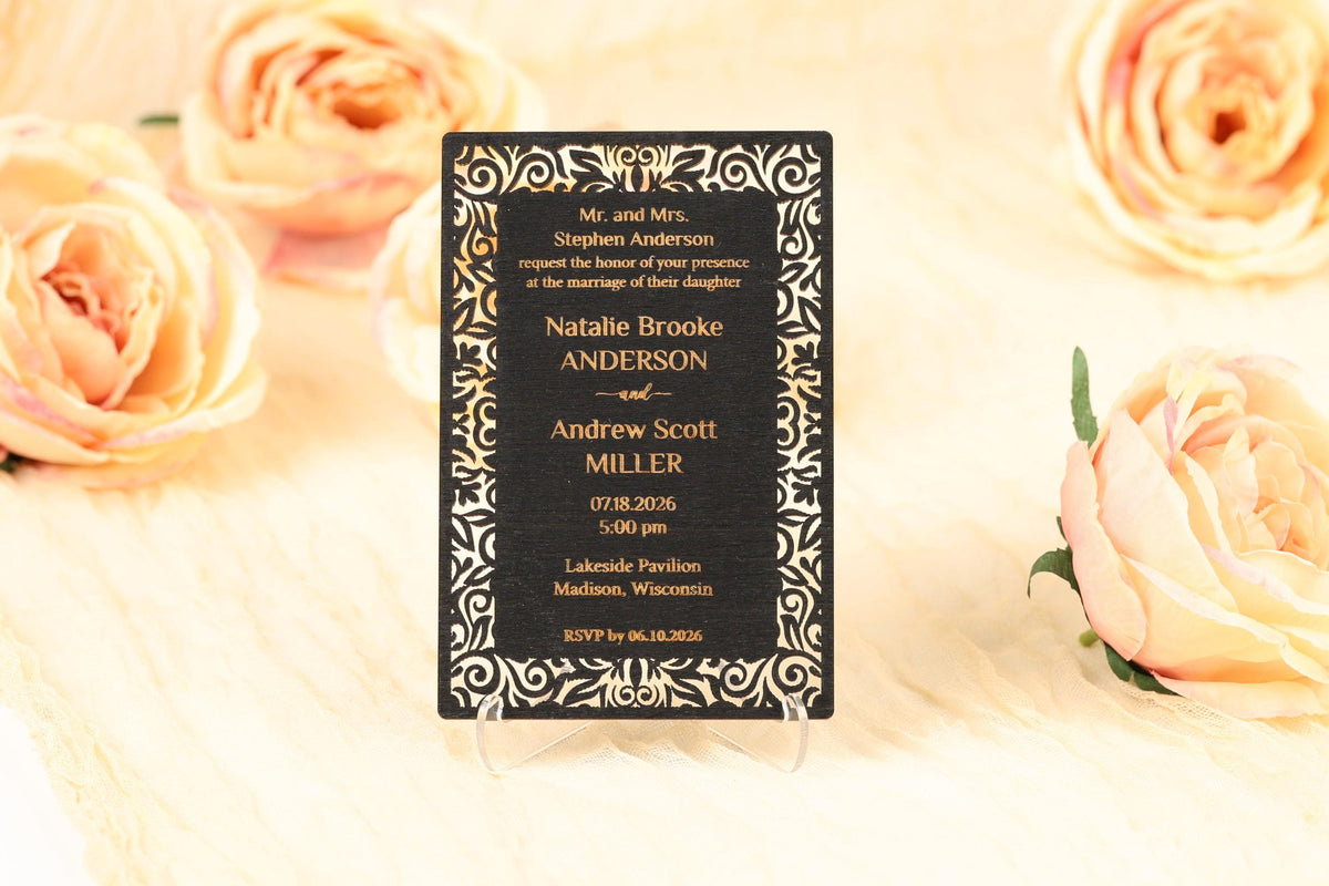 A black wedding invitation card with gold decorative borders, placed on a wooden surface with peach-colored roses in the background.