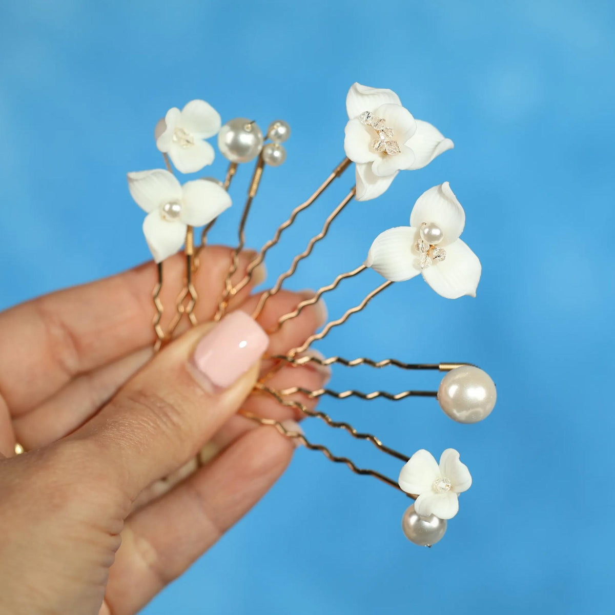 A hand holding a group of white flower-shaped hair clips with pearls attached against a blue background.