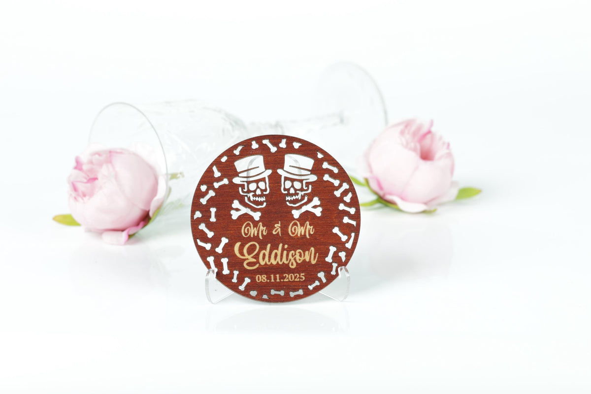 A wooden coaster with a circular design featuring two skull faces and the text "Mr & Mrs Eddison" is placed on a white surface, with two pink roses in the background.