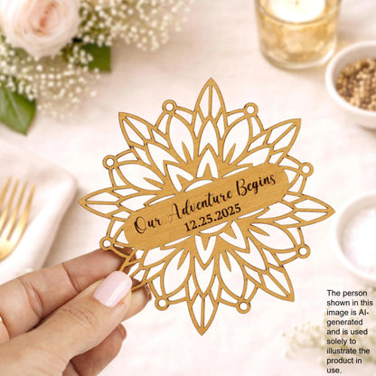 A hand holding a wooden ornament with the text "Our Adventure Begins 12.25.25" on it, surrounded by flowers and other decorative elements.