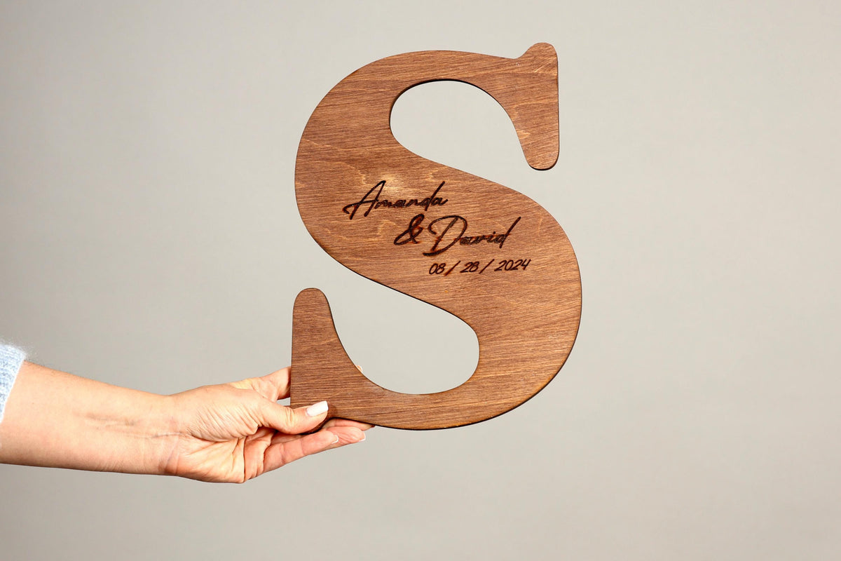 A person's hand holding a wooden letter 'S' with the names 'Amanda & David' engraved on it.