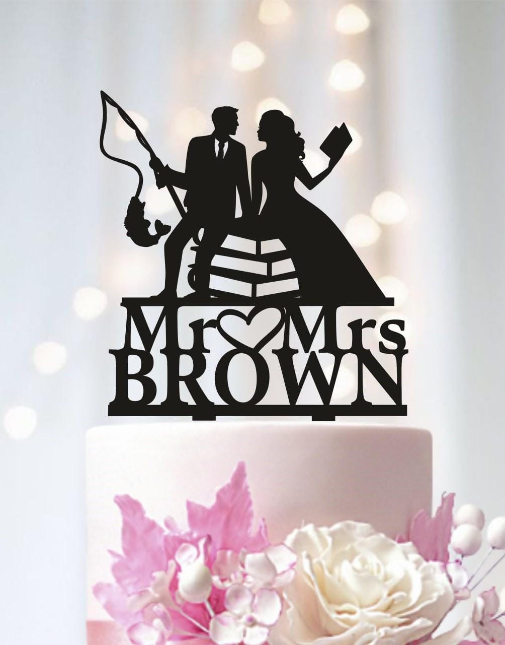 A black silhouette of a man and a woman holding a book and a fishing rod, standing on a cake topper that says "Mr & Mrs Brown".