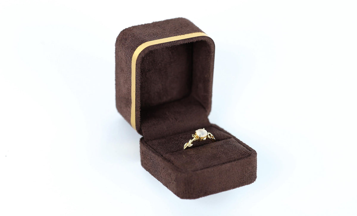 A gold ring with a diamond is displayed in a brown jewelry box against a white background.