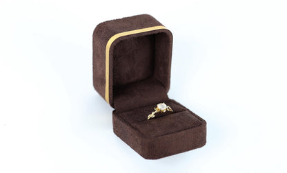 A gold ring with a diamond is displayed in a brown jewelry box against a white background.