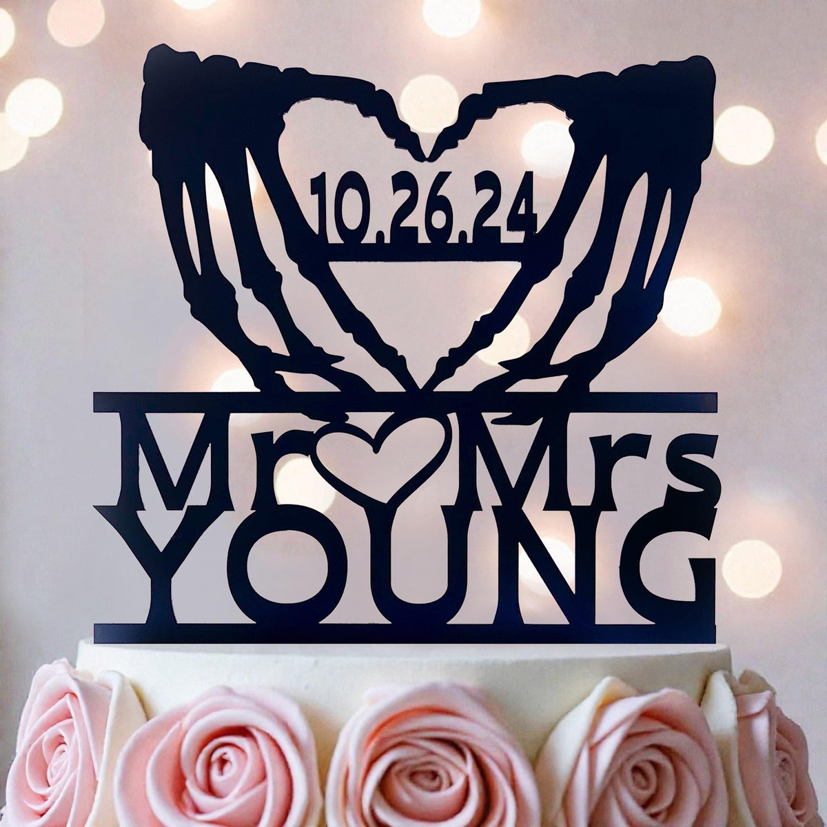 A black silhouette of a heart with the text "10.26.24" and "Mr. YOUNG" written on it, placed on top of a white cake decorated with pink roses.