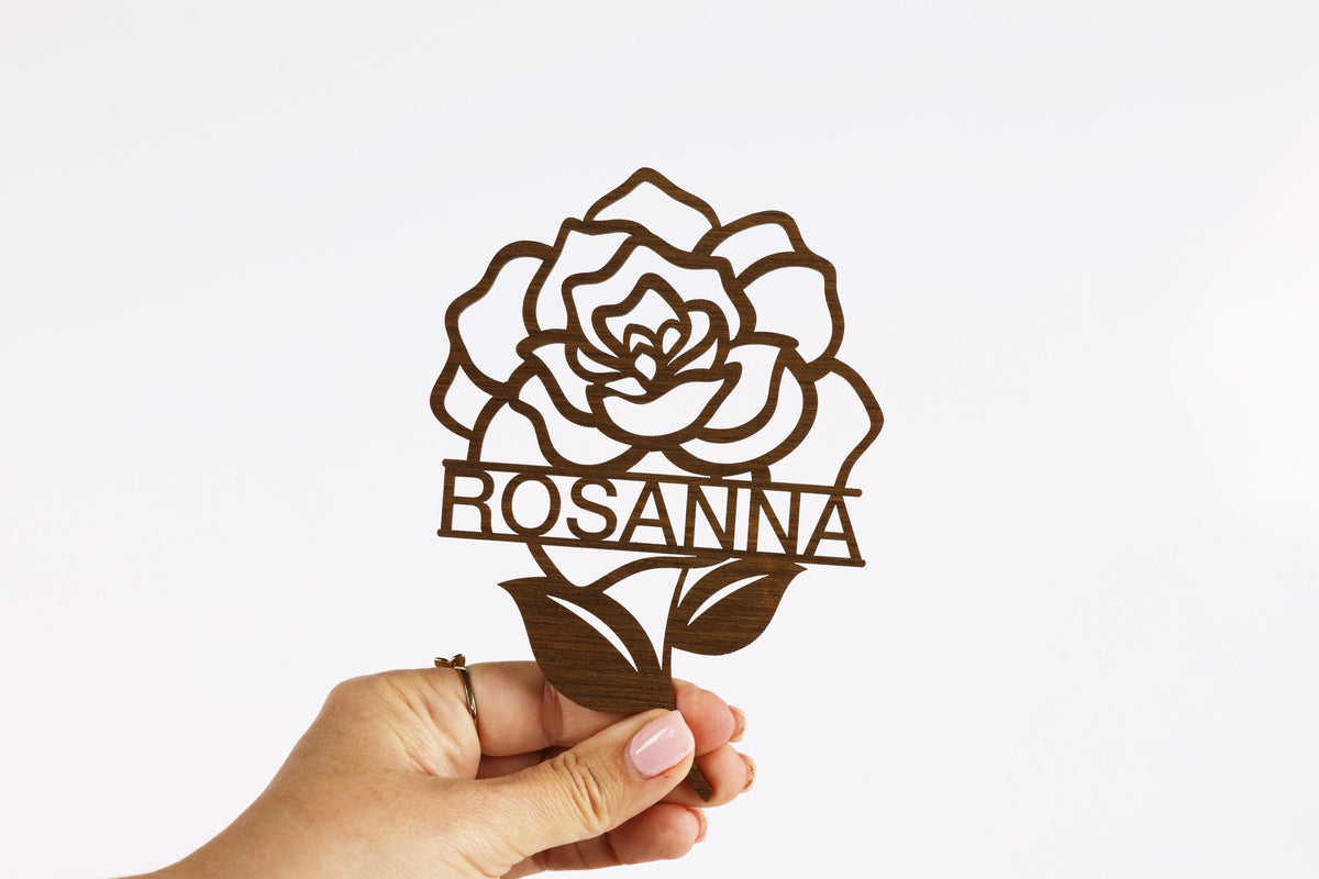 A hand holding a wooden cutout of a rose with the name "ROSANNA" written on it.