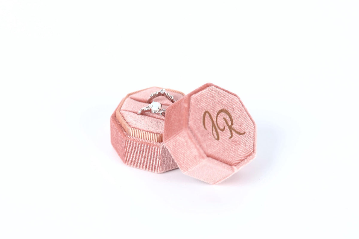 A pink velvet box containing a diamond ring, with the letters "JR" visible on the side of the box.