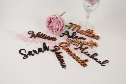 a collection of wooden name cards with names written on them, along with a pink rose and a wine glass.