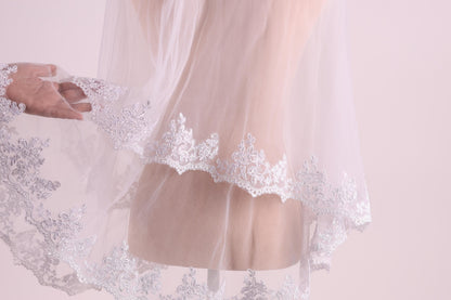 A close-up view of a delicate white lace veil with intricate floral embroidery, held by a hand.