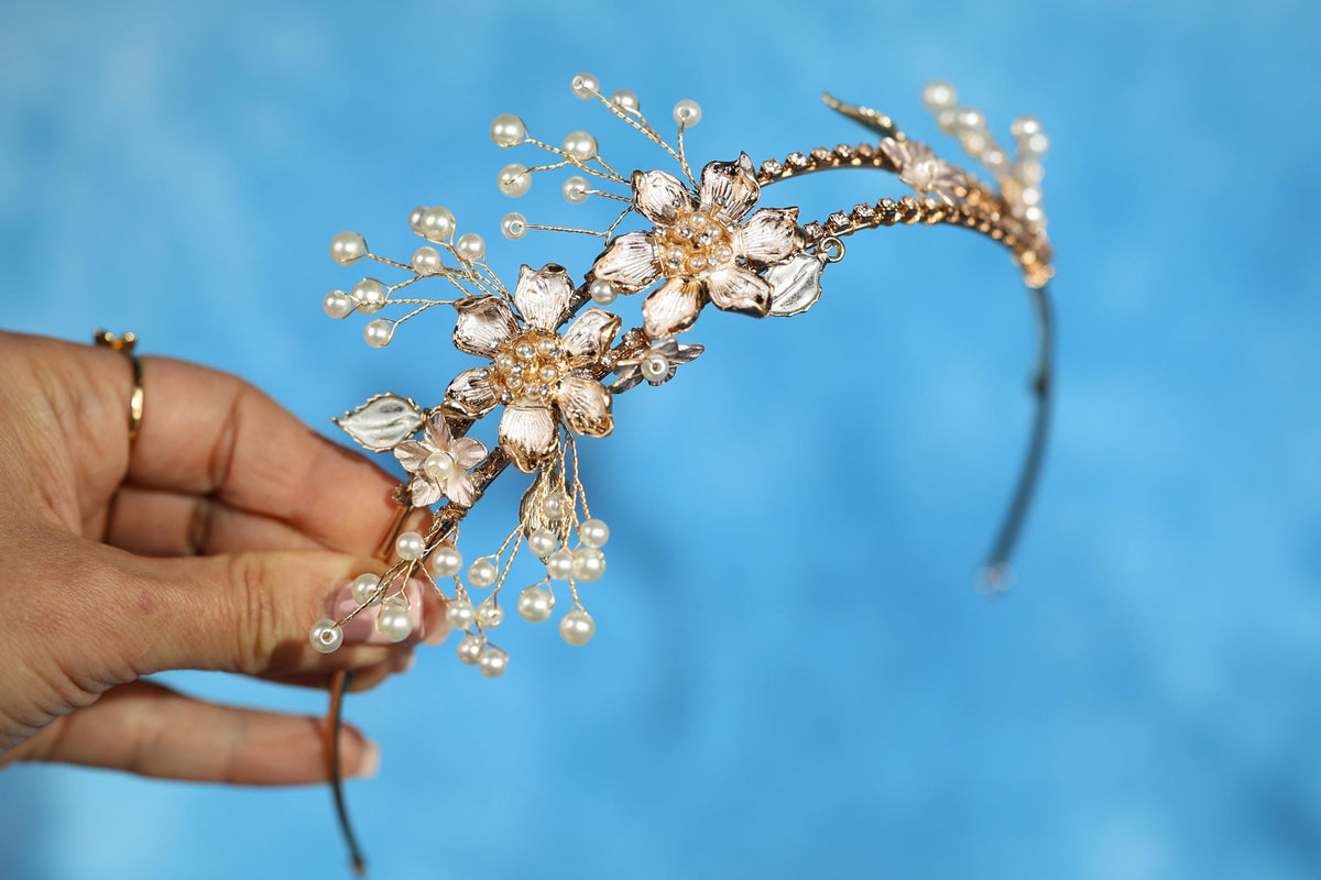 A hand holding a decorative headband with a floral design and small pearls against a blue background.