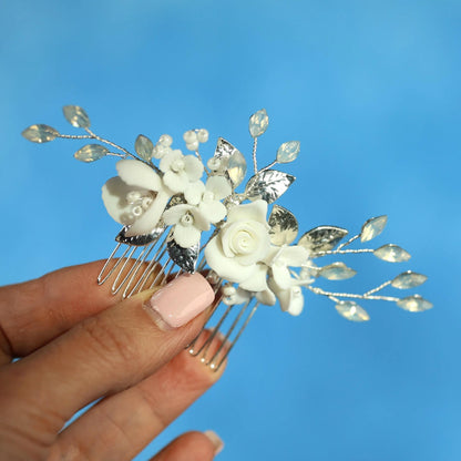 A hand holding a decorative hair clip with white flowers and leaves against a blue background.