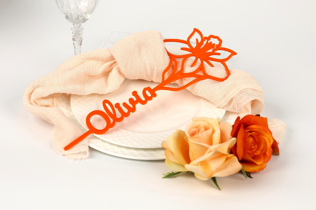 a table setting with a white plate, a glass, and a decorative orange flower. The table setting includes a napkin with the name "Olivia" written on it, and two roses are placed on the table.