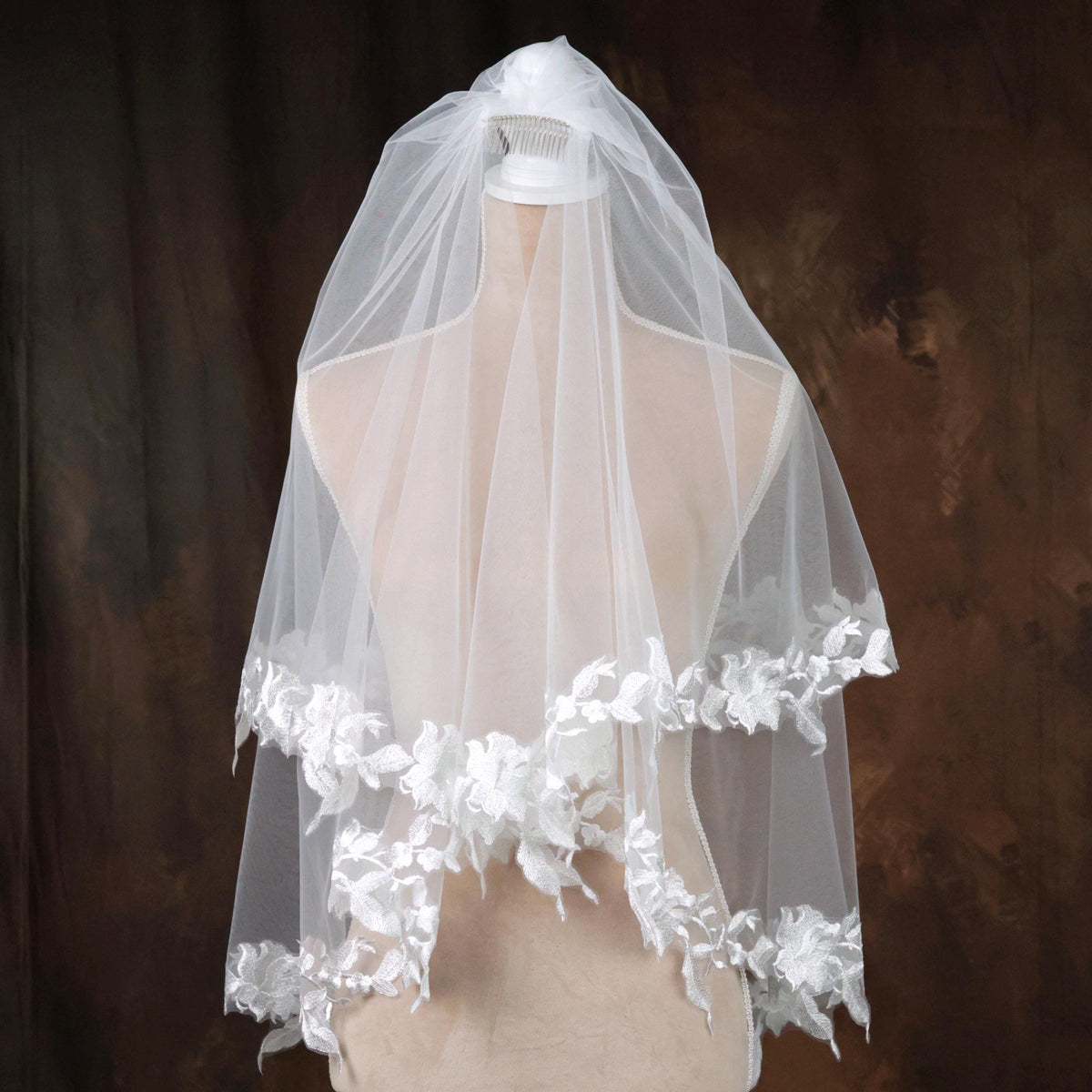 A white wedding veil with delicate white lace trim is displayed on a mannequin against a dark background.