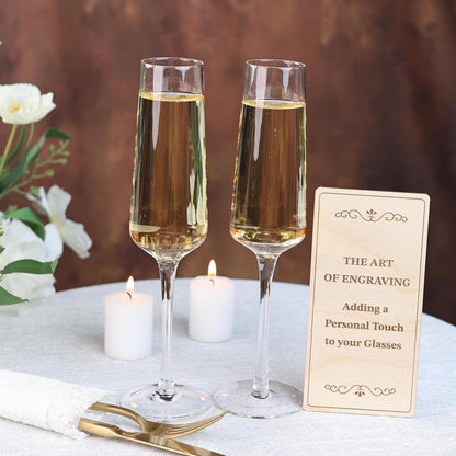 Two champagne glasses filled with golden liquid sit on a table, accompanied by a wooden sign that reads "The Art of Engraving: Adding a Personal Touch to your Glasses".
