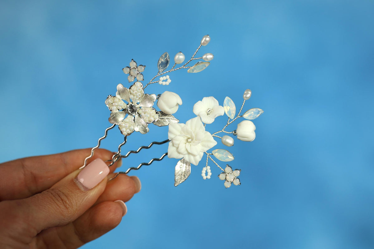 A hand holding a decorative hair clip with white flowers and leaves against a blue background.