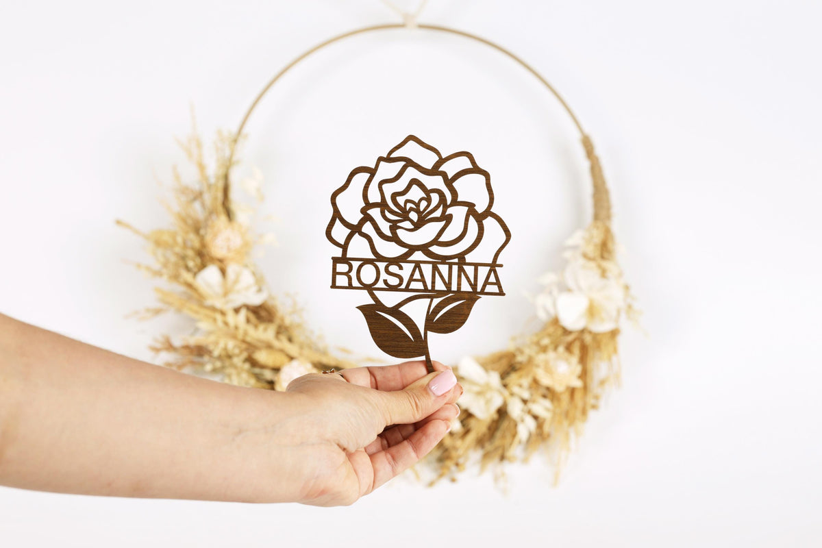 A hand holding a wooden rose-shaped cake topper with the name "ROSANNA" written on it.