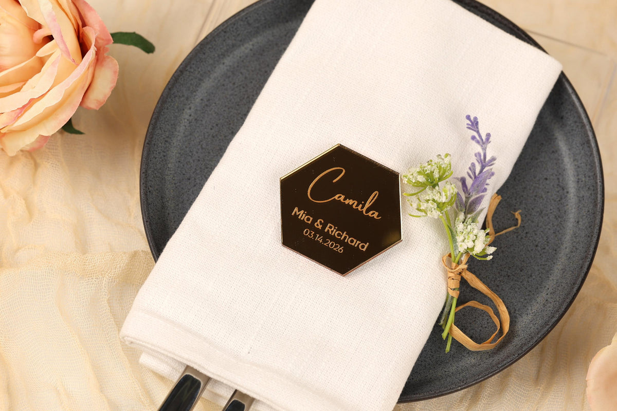 A black plate with a white napkin on it, featuring a small hexagonal card with the names 'Carmila' and 'Richard' on it, along with a floral arrangement.