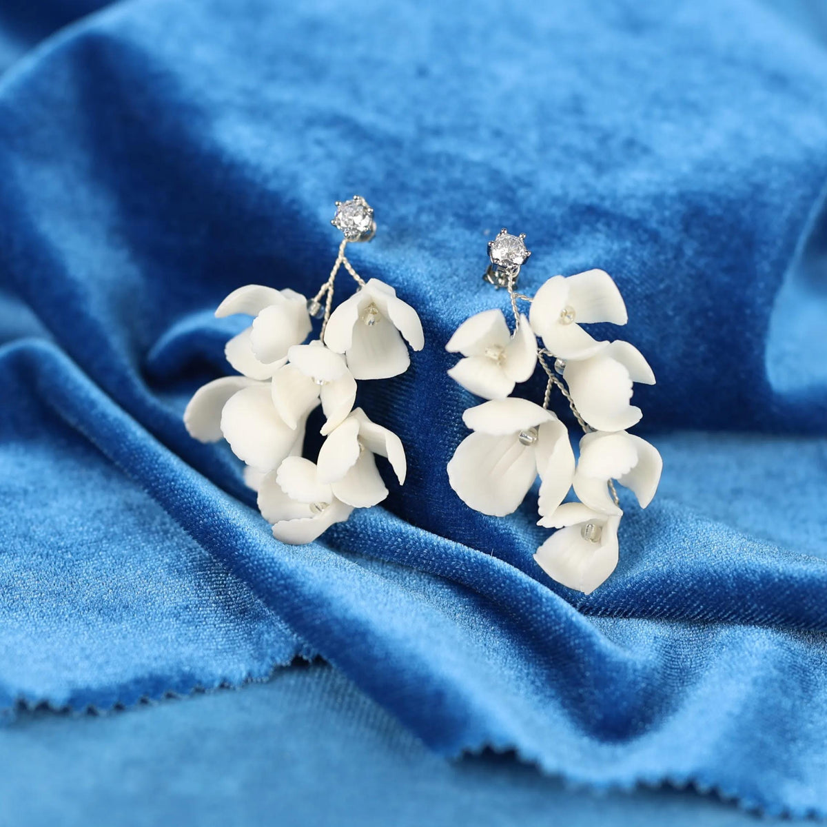 A pair of white flower-shaped earrings with small diamonds on top, resting on a blue fabric surface.