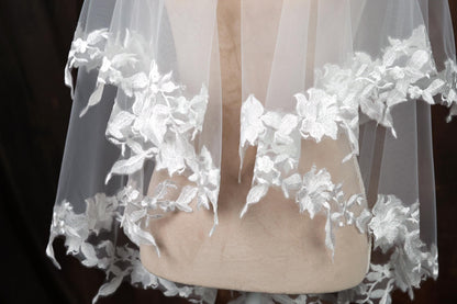 A close-up view of a delicate white lace veil with intricate floral embroidery, displayed on a mannequin or model.