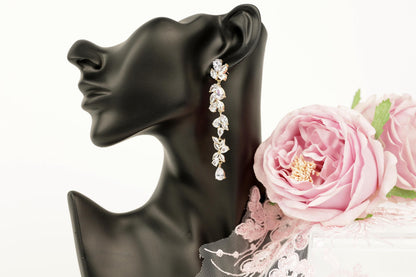 A black bust form with a pair of dangling earrings featuring a long, delicate chain design and small, clear gemstones. The earrings are placed next to a bouquet of pink roses, creating a romantic and elegant scene.