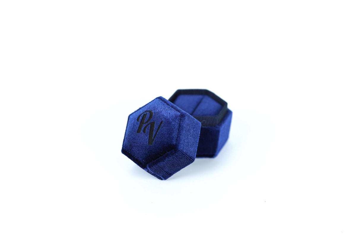 A blue velvet jewelry box with a hexagonal shape and a black logo on it.