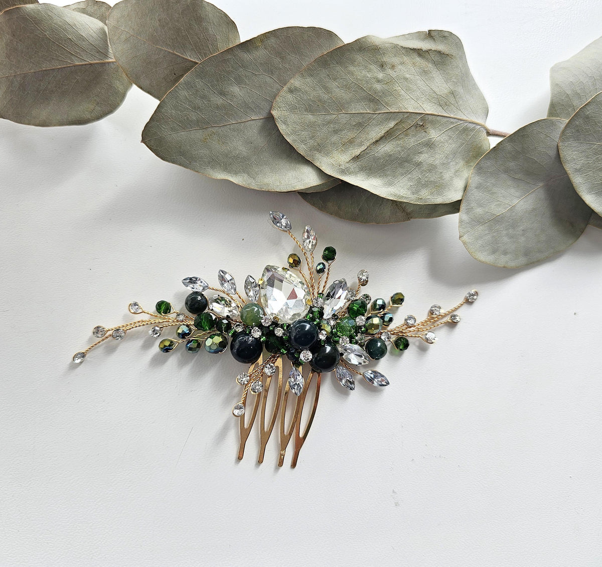 A decorative hair accessory with a gold-colored comb and a large crystal or gemstone at the center, surrounded by smaller stones and leaves.
