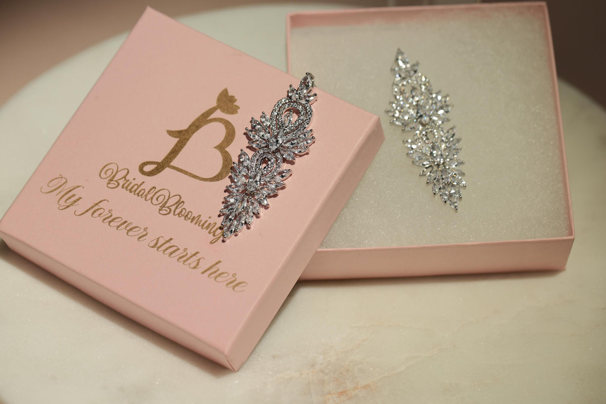 A pink jewelry box with a gold letter "B" and the text "My forever starts here" on it, along with a silver and white diamond earring placed inside the box.