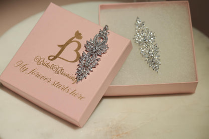 A pink jewelry box with a gold letter "B" and the text "My forever starts here" on it, along with a silver and white diamond earring placed inside the box.