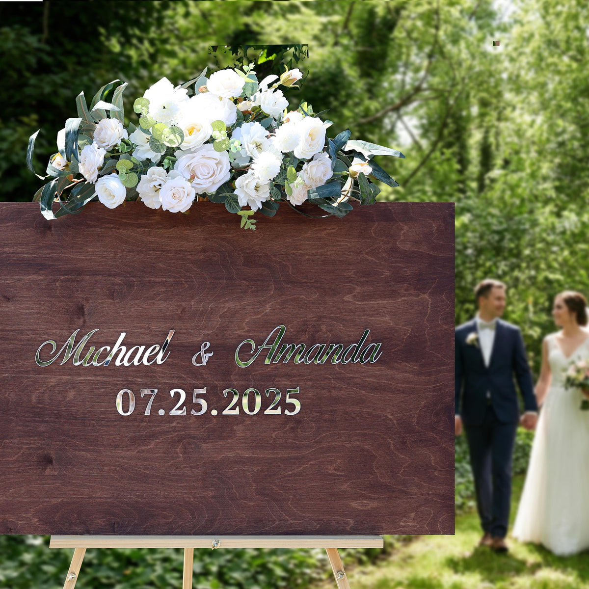 A wooden sign displaying the names Michael and Amanda, along with the date "07.25.2025", is prominently featured in the foreground. In the background, a couple dressed in wedding attire can be seen walking together.