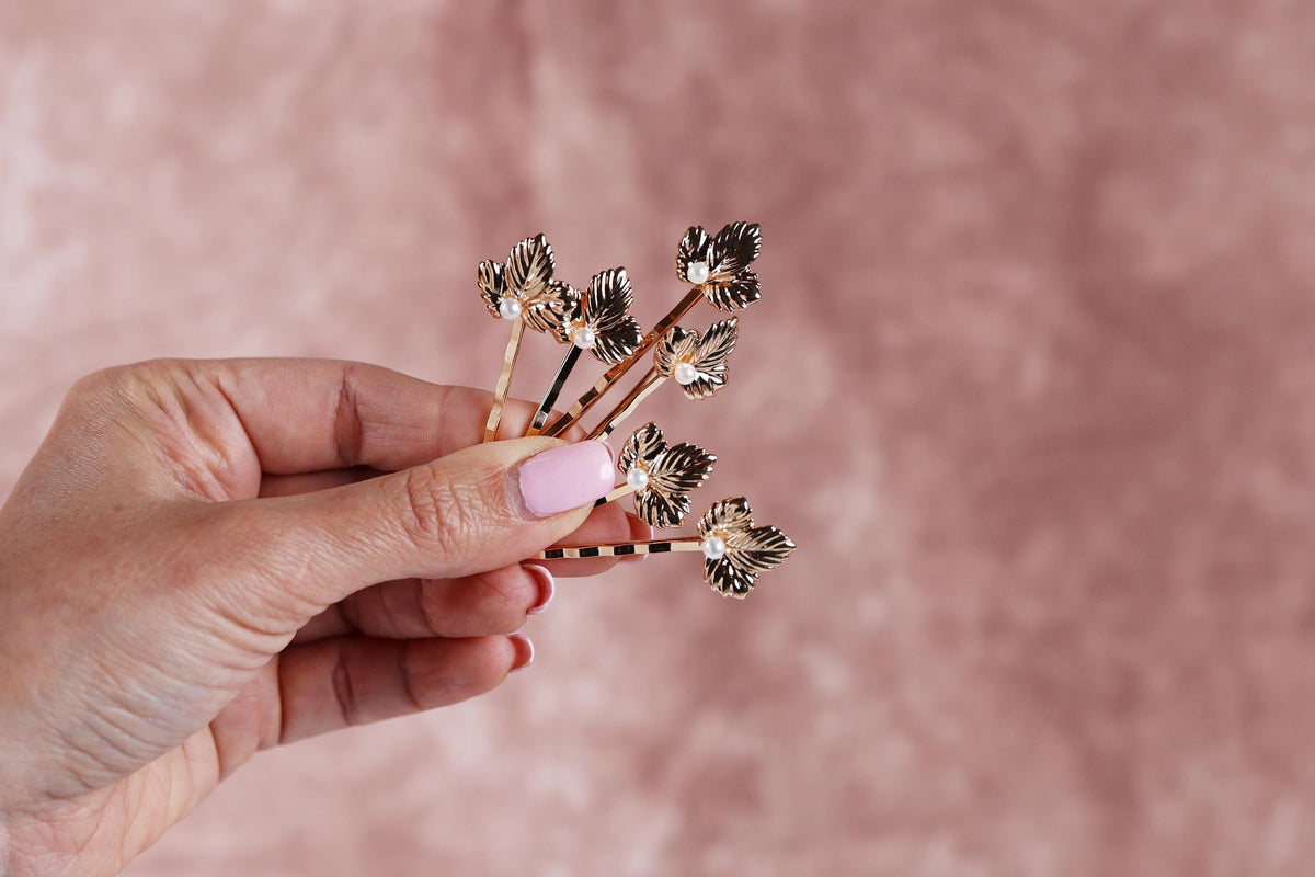 A hand holding a small group of gold-colored hair clips with leaf-shaped designs against a pink background.