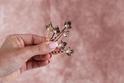 A hand holding a small group of gold-colored hair clips with leaf-shaped designs against a pink background.