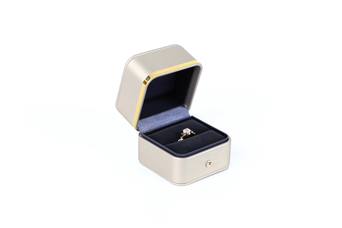 A gold ring is displayed in a gold and black jewelry box against a white background.