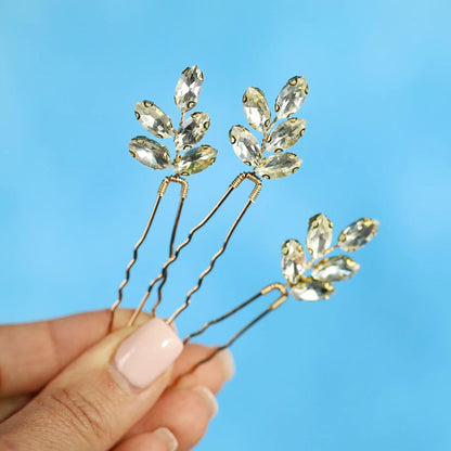 A hand holding three gold-colored hair clips with clear gemstone accents against a blue background.