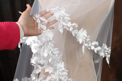 A hand is holding up a white veil with intricate silver lace designs.